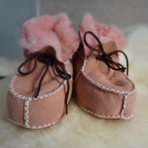 May include: A pair of baby booties in a soft pink suede. The booties have a fluffy pink fur trim around the ankle and are laced with brown cord. White stitching accents the edges of the shoe.