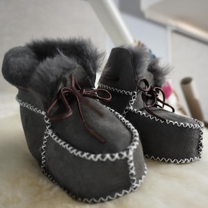 May include: A pair of gray suede booties with faux fur trim and white stitching. The booties have brown ribbon laces and are displayed on a white, fluffy surface. The style is reminiscent of moccasins.