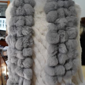 Puede incluir: Bufanda de piel gris con diseño de pompón, exhibida en un maniquí. La bufanda está hecha de piel suave y esponjosa y tiene una apariencia texturizada. La bufanda está drapeada sobre una prenda blanca, creando un contraste de color y textura.