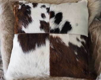Puede incluir: Una almohada cuadrada con un patrón de piel de vaca marrón y blanco. La almohada está hecha de cuatro piezas diferentes de piel de vaca, cada una con un patrón diferente. La almohada está sentada sobre una superficie de piel marrón claro.