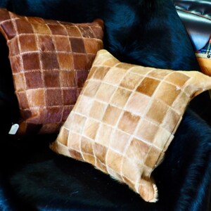 May include: Two square-shaped throw pillows with a patchwork design made of brown and tan leather. The pillows are sitting on a black fur throw.