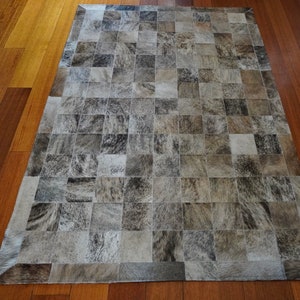 May include: A gray and white patchwork cowhide rug with a square pattern. The rug is made of individual pieces of cowhide that have been sewn together.