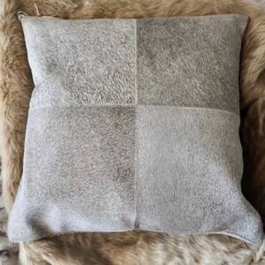 Puede incluir: Una almohada de piel de vaca gris y blanca con un patrón cuadrado. La almohada está hecha de cuero natural suave y tiene un brillo sutil.