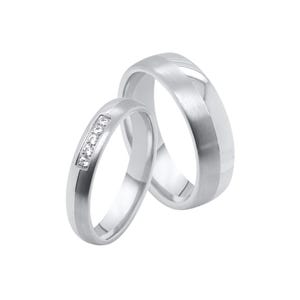Puede incluir: Dos anillos de boda plateados. Uno presenta una fila de pequeñas piedras transparentes. El otro tiene un acabado de metal cepillado con una banda central pulida. Ambos anillos están sobre un fondo blanco.