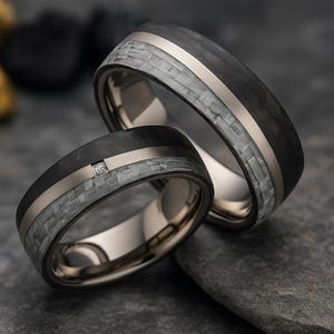 以下が含まれることがあります： 黒いカーボンファイバーバンド、シルバーのストライプ、グレーの織り模様が特徴の2つの結婚指輪。1つの指輪には小さなダイヤモンドが付いています。指輪は濃い灰色の表面に置かれています。