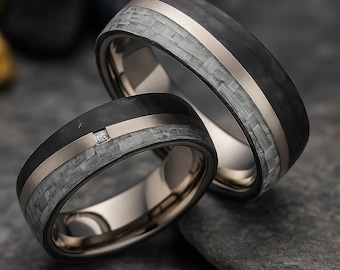 May include: Two wedding rings with a black carbon fiber band, a silver stripe, and a gray woven pattern. One ring has a small diamond. The rings are on a dark gray surface.
