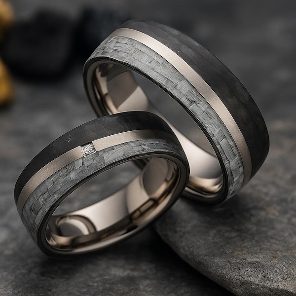 May include: Two wedding rings with a black carbon fiber band, a silver stripe, and a gray woven pattern. One ring has a small diamond. The rings are on a dark gray surface.