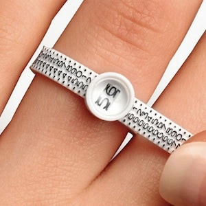 Könnte beinhalten: Ein weißer Kunststoff-Ringgrößenmesser mit schwarzen Markierungen und einem kleinen weißen Zifferblatt, auf dem "10" steht.