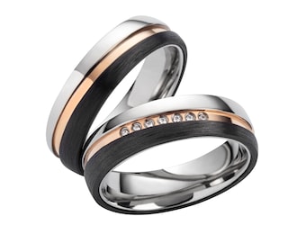 May include: Two wedding bands with a black carbon fiber inlay. One band has a rose gold stripe and the other has a row of small diamonds.