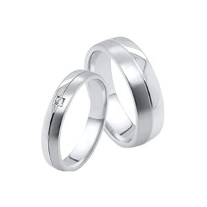 Puede incluir: Dos anillos de boda plateados. Uno tiene un pequeño diamante cuadrado y una ranura central. El otro tiene un acabado cepillado y un diseño de ranura similar. Ambos anillos están sobre un fondo blanco.