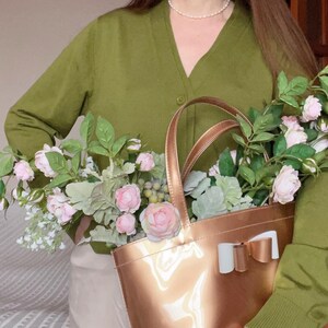 May include: A shiny, rose gold tote bag with a white and gold bow detail, filled with pink and white flowers and greenery. The bag is held in front of a person wearing a green cardigan.