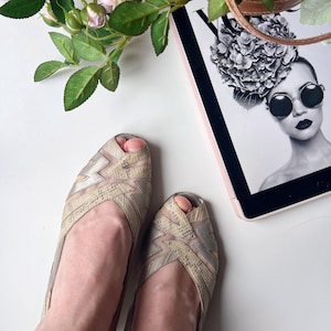 May include: A pair of open-toe flats with a geometric design in silver and beige. The shoes have a mesh overlay and a snakeskin-like pattern. The image also includes a tablet with a fashion magazine cover, pink roses, and a decorative light.