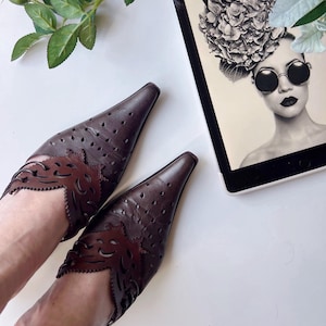 May include: A pair of dark brown leather pointed-toe flats with decorative cut-out detailing. The shoes are placed on a white surface next to a tablet displaying a black and white image of a woman with sunglasses and a floral headpiece.