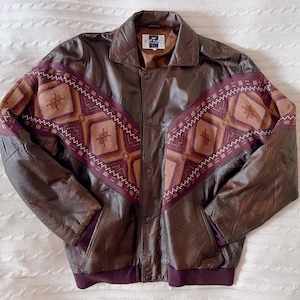 Puede incluir: Chaqueta de cuero marrón con un diseño geométrico en las mangas y la parte superior del pecho. La chaqueta tiene una parte delantera abotonada, un cuello y un dobladillo y puños acanalados. El patrón geométrico presenta tonos de marrón, beige y burdeos.