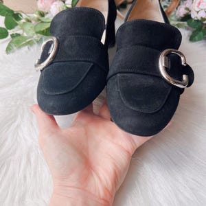 K&ouml;nnte beinhalten: Ein Paar schwarze Wildleder-Loafer mit abgerundeter Zehenpartie und einer markanten silberfarbenen Metallschnalle &uuml;ber dem Vorderblatt. Die Schuhe haben ein glattes Innenfutter und einen niedrigen Blockabsatz mit sichtbaren N&auml;hten.