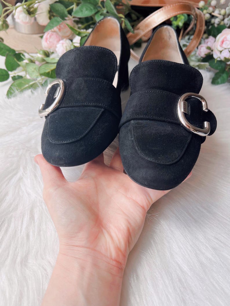 K&ouml;nnte beinhalten: Ein Paar schwarze Wildleder-Loafer mit abgerundeter Zehenpartie und einer markanten silberfarbenen Metallschnalle &uuml;ber dem Vorderblatt. Die Schuhe haben ein glattes Innenfutter und einen niedrigen Blockabsatz mit sichtbaren N&auml;hten.