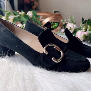 K&ouml;nnte beinhalten: Ein Paar schwarze Wildleder-Loafer mit niedrigem Blockabsatz und abgerundeter Schuhspitze. Jeder Schuh ist mit einer auff&auml;lligen, polierten silberfarbenen Metallschnalle auf dem Vorderblatt verziert, die ein kleines eingraviertes Logo-Detail tr&auml;gt.