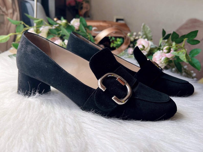 K&ouml;nnte beinhalten: Ein Paar schwarze Wildleder-Loafer mit niedrigem Blockabsatz und abgerundeter Schuhspitze. Jeder Schuh ist mit einer auff&auml;lligen, polierten silberfarbenen Metallschnalle auf dem Vorderblatt verziert, die ein kleines eingraviertes Logo-Detail tr&auml;gt.