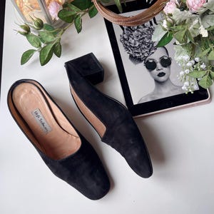 May include: Black suede slip-on shoes with a low heel, displayed on a white surface. The image also shows a black box, a tablet with a monochrome image, and floral decorations. The shoes have the text "Stefi Talman".
