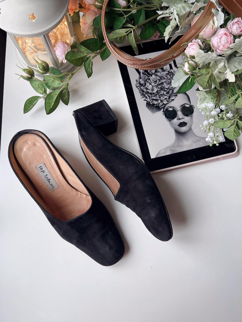 May include: Black suede slip-on shoes with a low heel, displayed on a white surface. The image also shows a black box, a tablet with a monochrome image, and floral decorations. The shoes have the text "Stefi Talman".