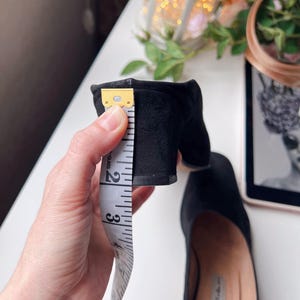 May include: Black suede block heel shoe with a heel height of approximately 6.35 cm. The shoe is being measured with a white measuring tape. The shoe is on a white surface with a blurred background.