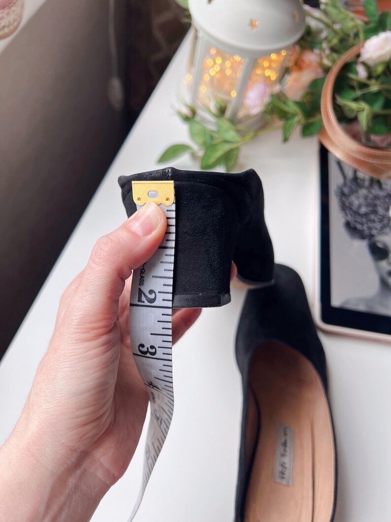 May include: Black suede block heel shoe with a heel height of approximately 6.35 cm. The shoe is being measured with a white measuring tape. The shoe is on a white surface with a blurred background.