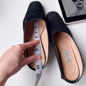 May include: Black suede slip-on shoes with a rounded toe. A measuring tape shows the shoe's interior length. The shoes are on a white surface. The shoes have a label that reads "Seymour, 4725".