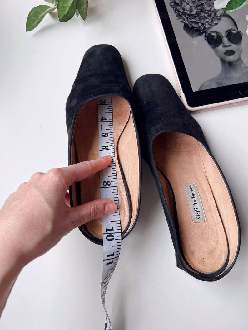 May include: Black suede slip-on shoes with a rounded toe. A measuring tape shows the shoe's interior length. The shoes are on a white surface. The shoes have a label that reads "Seymour, 4725".