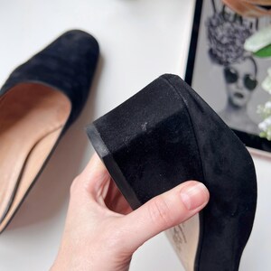 May include: Black suede block heel shoe. The shoe has a rounded toe and a low, sturdy heel. The sole is a light tan colour. The shoe is held by a hand, showing the heel and sole. The other shoe is visible in the background.