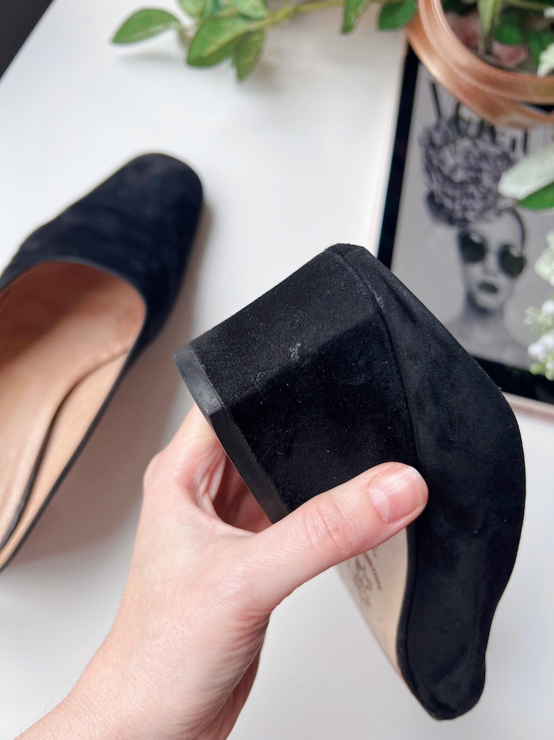 May include: Black suede block heel shoe. The shoe has a rounded toe and a low, sturdy heel. The sole is a light tan colour. The shoe is held by a hand, showing the heel and sole. The other shoe is visible in the background.