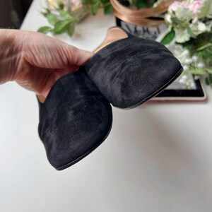 May include: A pair of black suede slip-on shoes with a square toe. The shoes are held by a hand, showcasing the soft texture of the material. The background includes a white surface and decorative elements.