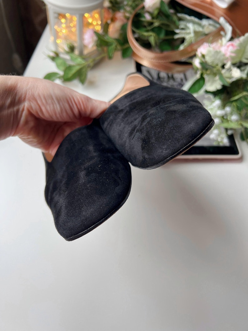 May include: A pair of black suede slip-on shoes with a square toe. The shoes are held by a hand, showcasing the soft texture of the material. The background includes a white surface and decorative elements.