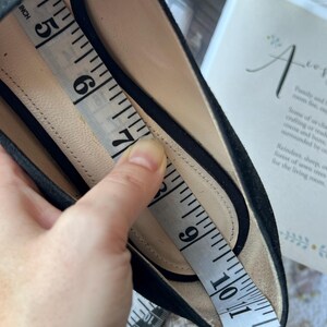 K&ouml;nnte beinhalten: Ein schwarzer Schuh mit beigem Innenfutter. Ein wei&szlig;es Ma&szlig;band liegt im Schuh und zeigt eine L&auml;ngenmessung von etwa 12,7 cm bis 25,4 cm entlang der Innensohle des Schuhwerks an.