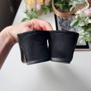 May include: Close-up of a pair of black suede slip-on mules. The shoes feature a rounded toe and a small block heel. The image highlights the suede texture and the heel's shape. The mules are a classic style.