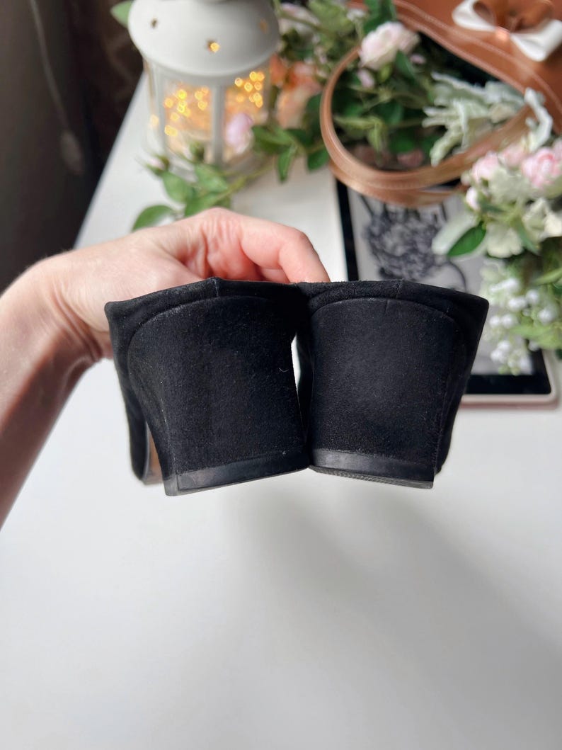 May include: Close-up of a pair of black suede slip-on mules. The shoes feature a rounded toe and a small block heel. The image highlights the suede texture and the heel's shape. The mules are a classic style.