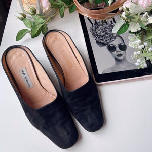 May include: Black suede slip-on mules with a rounded toe. The shoes have a light brown insole and a small label with the text "Stell Tolman". The shoes are on a white surface next to a tablet with a black and white image.