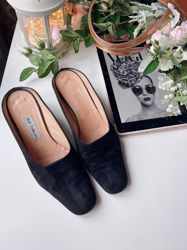 May include: Black suede slip-on mules with a rounded toe. The shoes have a light brown insole and a small label with the text "Stell Tolman". The shoes are on a white surface next to a tablet with a black and white image.