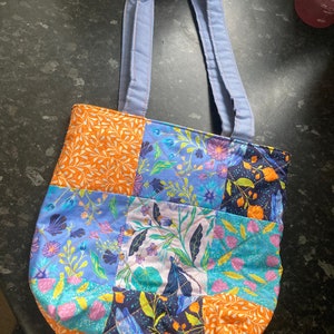 May include: A blue quilted tote bag with a patchwork design featuring floral and geometric patterns in shades of orange, blue, green, and pink.