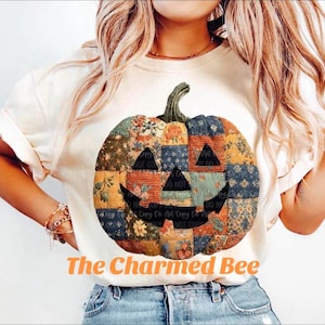 May include: Cream-colored t-shirt featuring a patchwork pumpkin design with a smiling jack-o'-lantern face. The pumpkin is made of various floral and patterned fabrics. The text "The Charmed Bee" is printed in orange below the pumpkin.