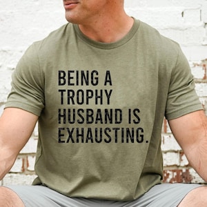 May include: A green t-shirt with the text "Being a Trophy Husband is Exhausting."