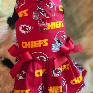 Puede incluir: Vestido rojo para perro con un patr&oacute;n repetido de cascos de f&uacute;tbol, logotipos del equipo y la palabra "CHIEFS" en amarillo. El vestido tiene una falda con volantes y est&aacute; adornado con lazos rojos.