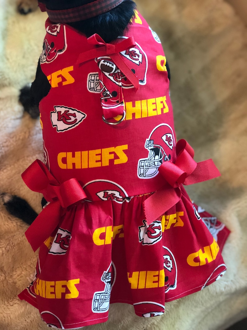 Puede incluir: Vestido rojo para perro con un patr&oacute;n repetido de cascos de f&uacute;tbol, logotipos del equipo y la palabra "CHIEFS" en amarillo. El vestido tiene una falda con volantes y est&aacute; adornado con lazos rojos.