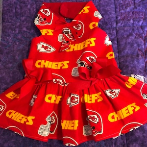 Puede incluir: Vestido rojo para perro con el logotipo y el casco de los Kansas City Chiefs. El vestido tiene una falda con volantes, un lazo en la cintura y un cuello. La palabra "CHIEFS" est&aacute; impresa en amarillo.