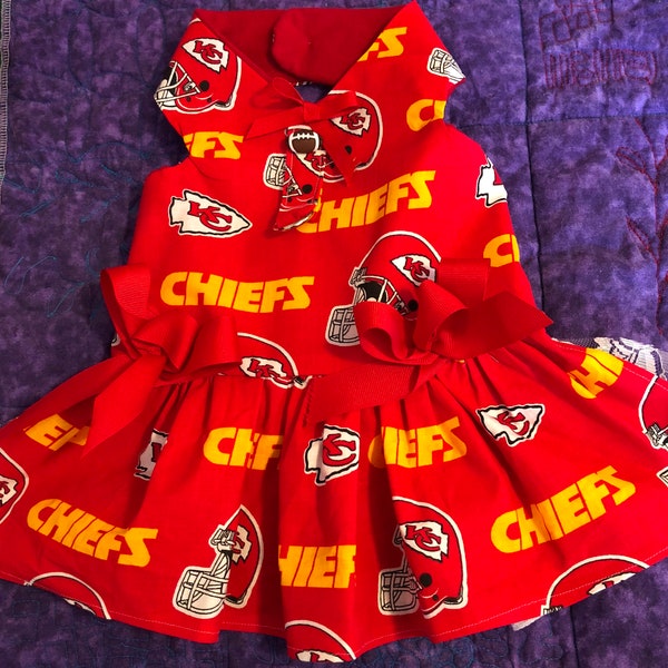 May include: A red dog dress featuring the Kansas City Chiefs logo and helmet design. The dress has a ruffled skirt, a bow at the waist, and a collar. The word "CHIEFS" is printed in yellow.