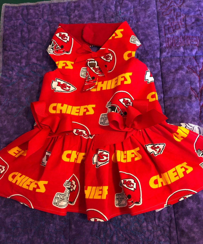 Puede incluir: Vestido rojo para perro con el logotipo y el casco de los Kansas City Chiefs. El vestido tiene una falda con volantes, un lazo en la cintura y un cuello. La palabra "CHIEFS" est&aacute; impresa en amarillo.