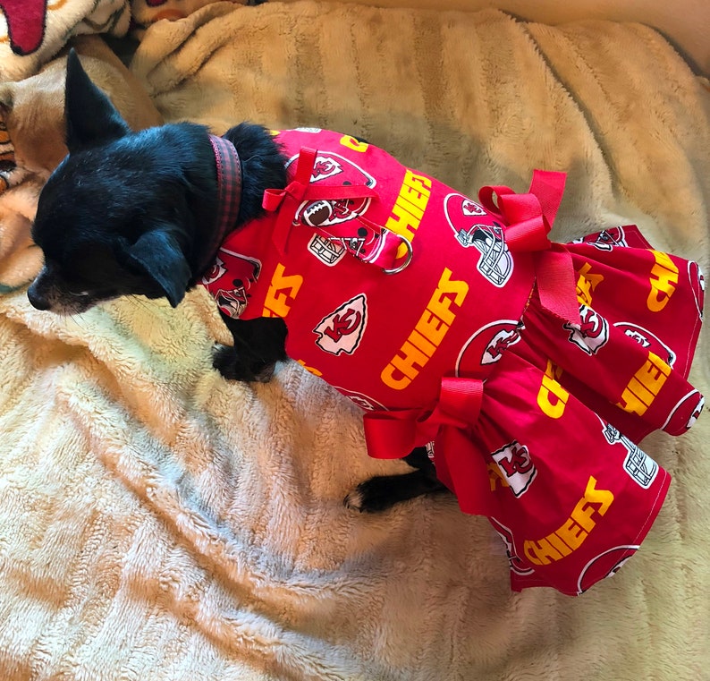 Puede incluir: Un perro peque&ntilde;o y negro con un vestido rojo y amarillo de los Kansas City Chiefs. El vestido presenta el logotipo de los Chiefs y la palabra "CHIEFS" en amarillo. El vestido tiene lazos rojos y una falda con volantes.