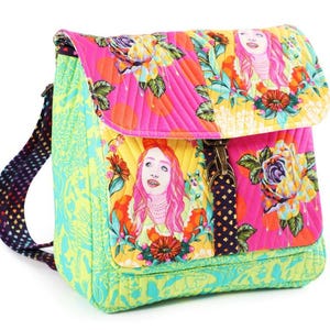 Puede incluir: Una mochila de tela acolchada con un vibrante estampado floral multicolor e ilustraciones de una persona con cabello rosa. La bolsa incluye un panel lateral verde lima, una correa oscura con un patr&oacute;n peque&ntilde;o y un cierre de metal en el frente.