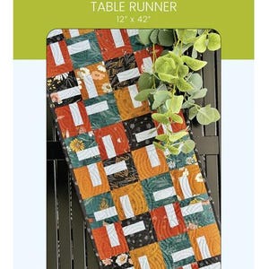 May include: A colorful table runner with a patchwork design. The runner is 12 inches by 42 inches and features squares in orange, green, black, and white with floral and geometric patterns. The text "Lil' Inga" and "Table Runner" are visible.