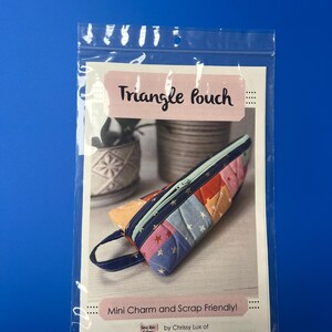 May include: A colorful, patchwork triangle pouch with a navy blue zipper and handle. The pouch is displayed inside a clear plastic bag with the text "Triangle Pouch" and "Mini Charm and Scrap Friendly!"