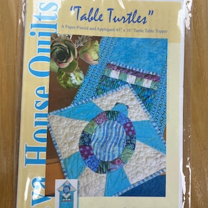 May include: A paper-pieced and appliquéd quilt pattern titled "Table Turtles" for a 45" x 16" turtle table topper. The pattern features a colorful turtle design with blue, white, and purple hues. The package is sealed in a clear plastic bag.